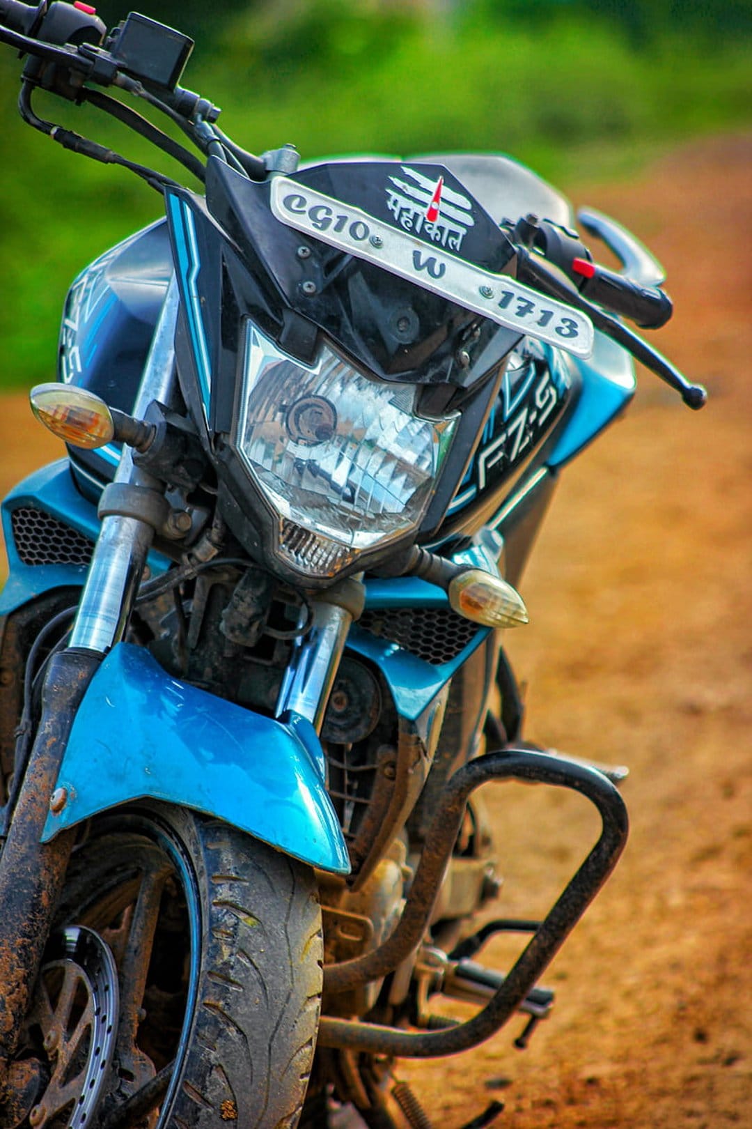 Blue Yamaha FZ-S V2 motorcycle with a sharp headlight parked on a gravel road against a blurred green background.