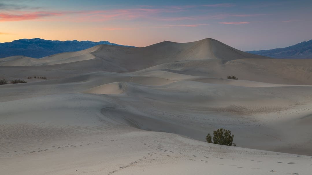 Towering sand dunes feature sharp ridges and ripple patterns under a soft pastel twilight sky in the desert.
