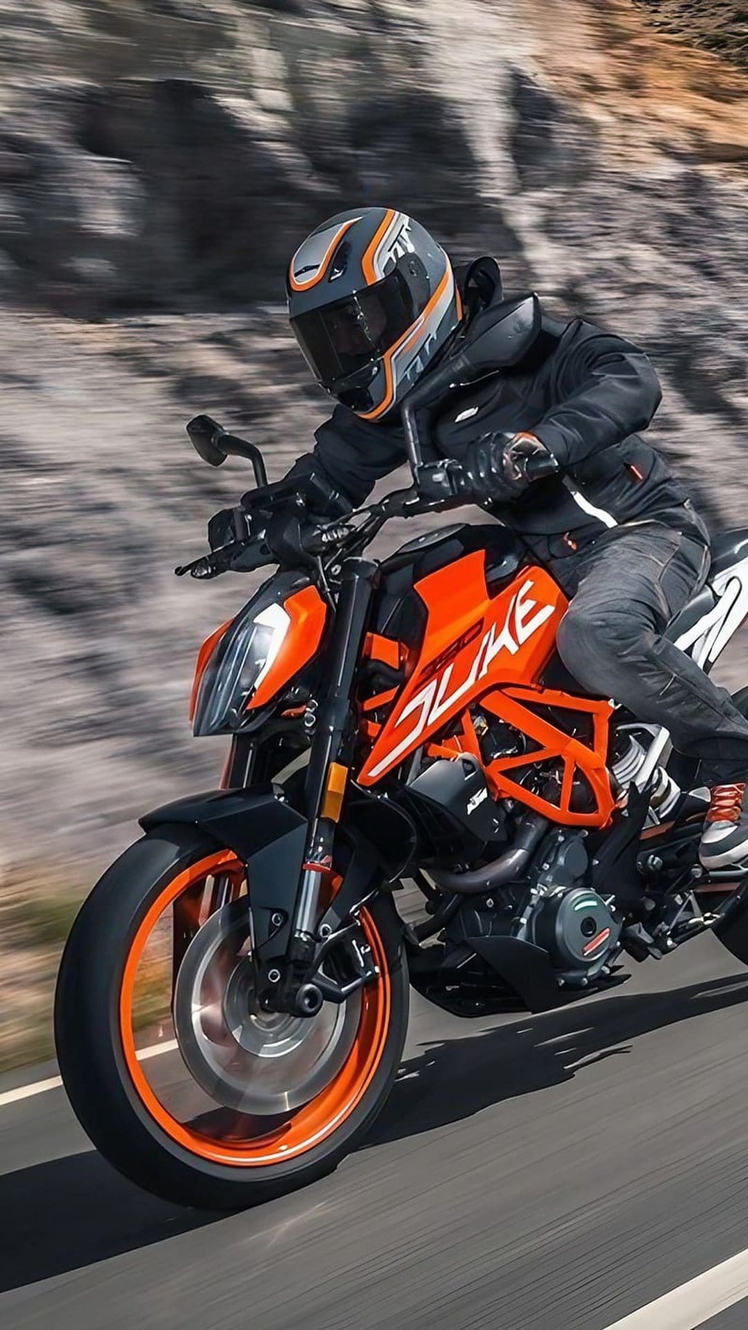 Motorcyclist in black gear rides an orange KTM Duke on a paved road with motion blur and a rocky background.