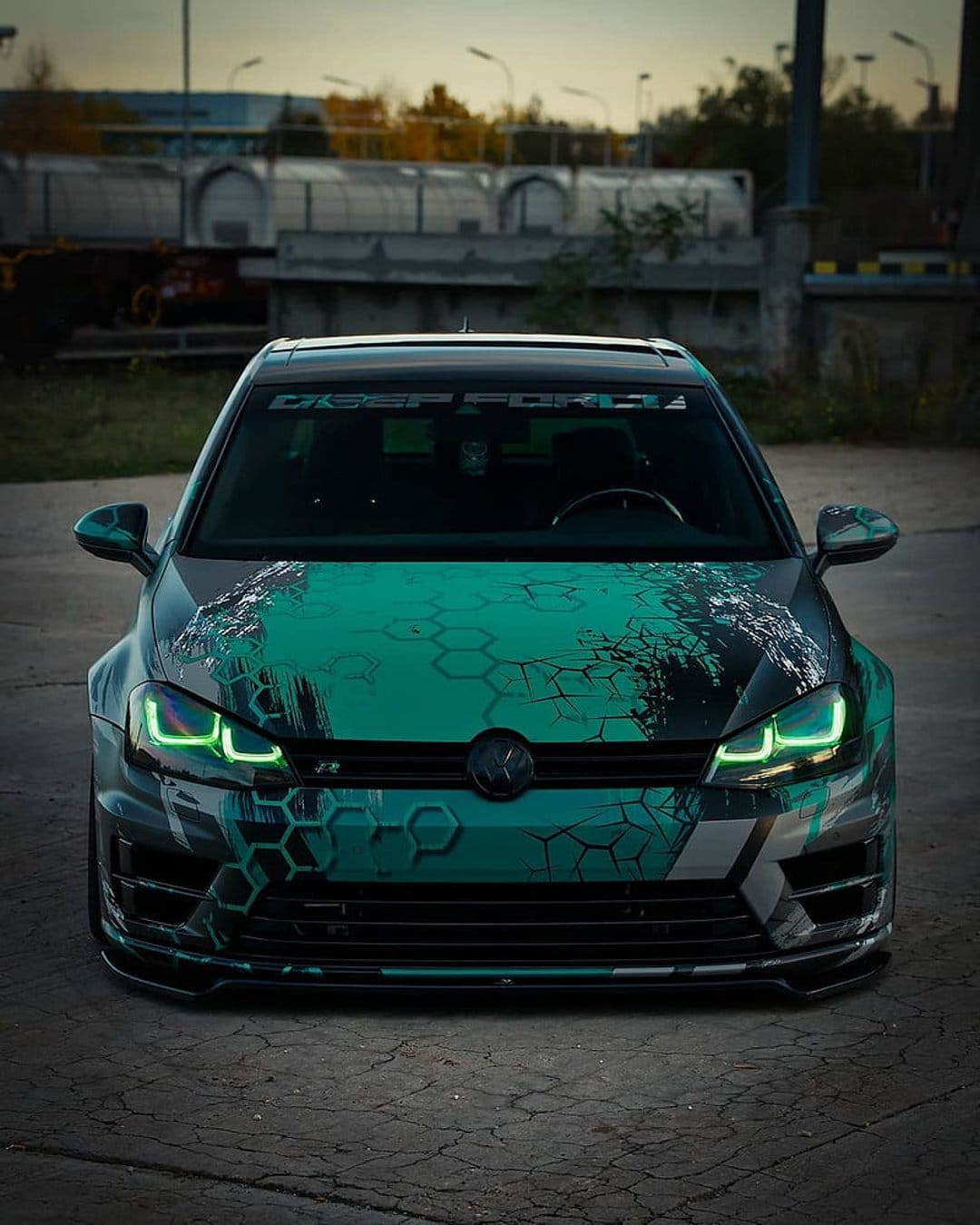 Volkswagen Golf R with a teal and black hexagonal wrap parked on concrete during a warm golden hour sunset.