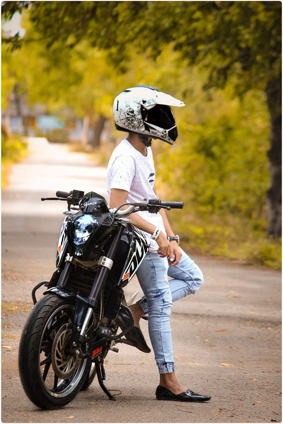 Man in a motorcycle helmet and white t-shirt poses with a bike on a road surrounded by autumn trees.