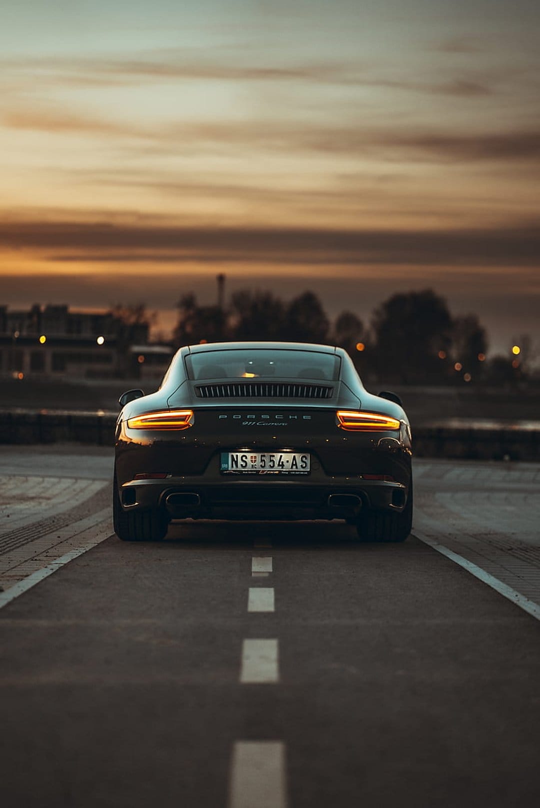 Porsche 911 Carrera rear view on an asphalt road at sunset with glowing red taillights and a cloudy orange sky.