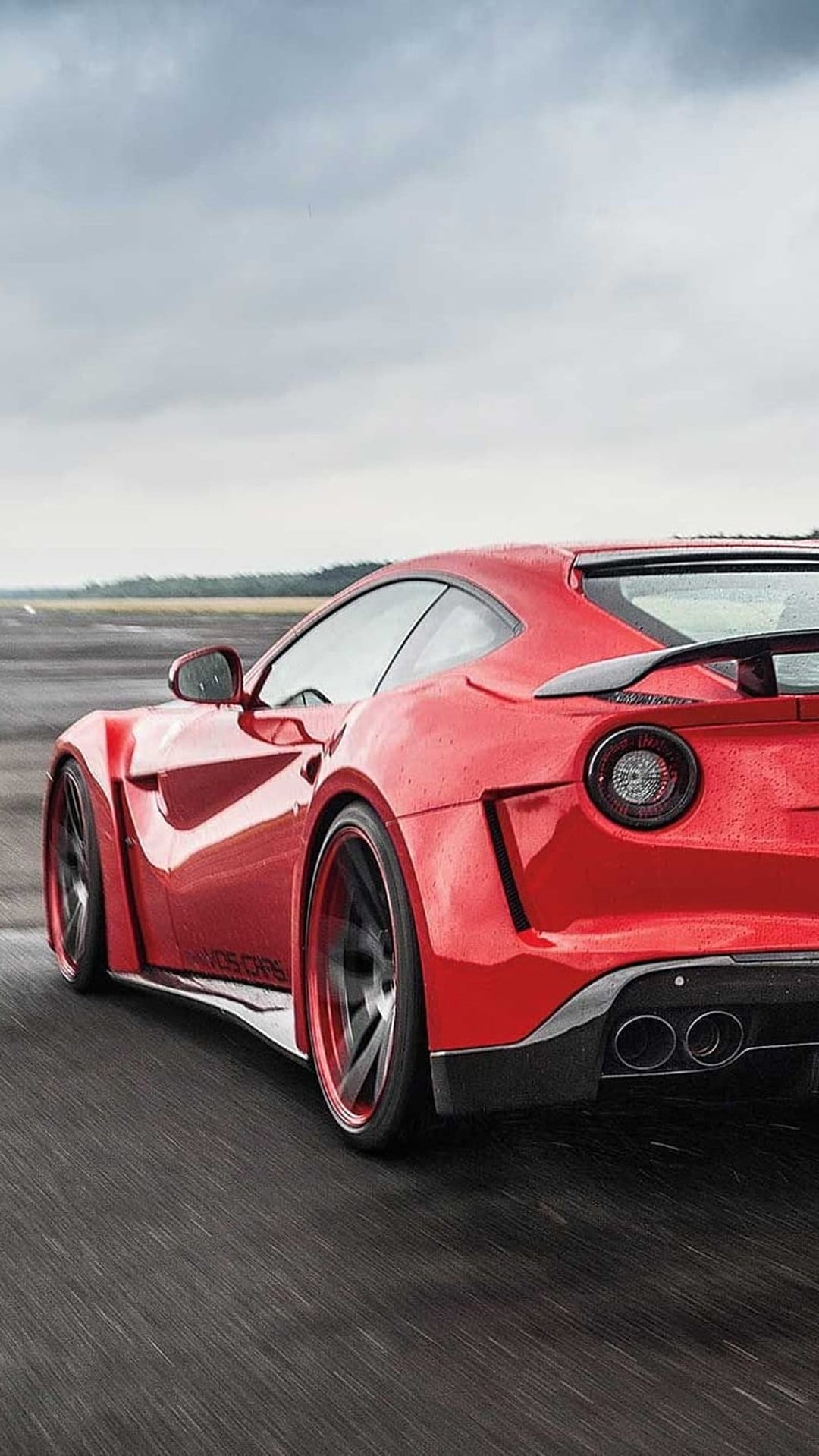 Red Ferrari F12tdf speeding on a wet racetrack with motion blur, wide body kit, and a cloudy sky backdrop.