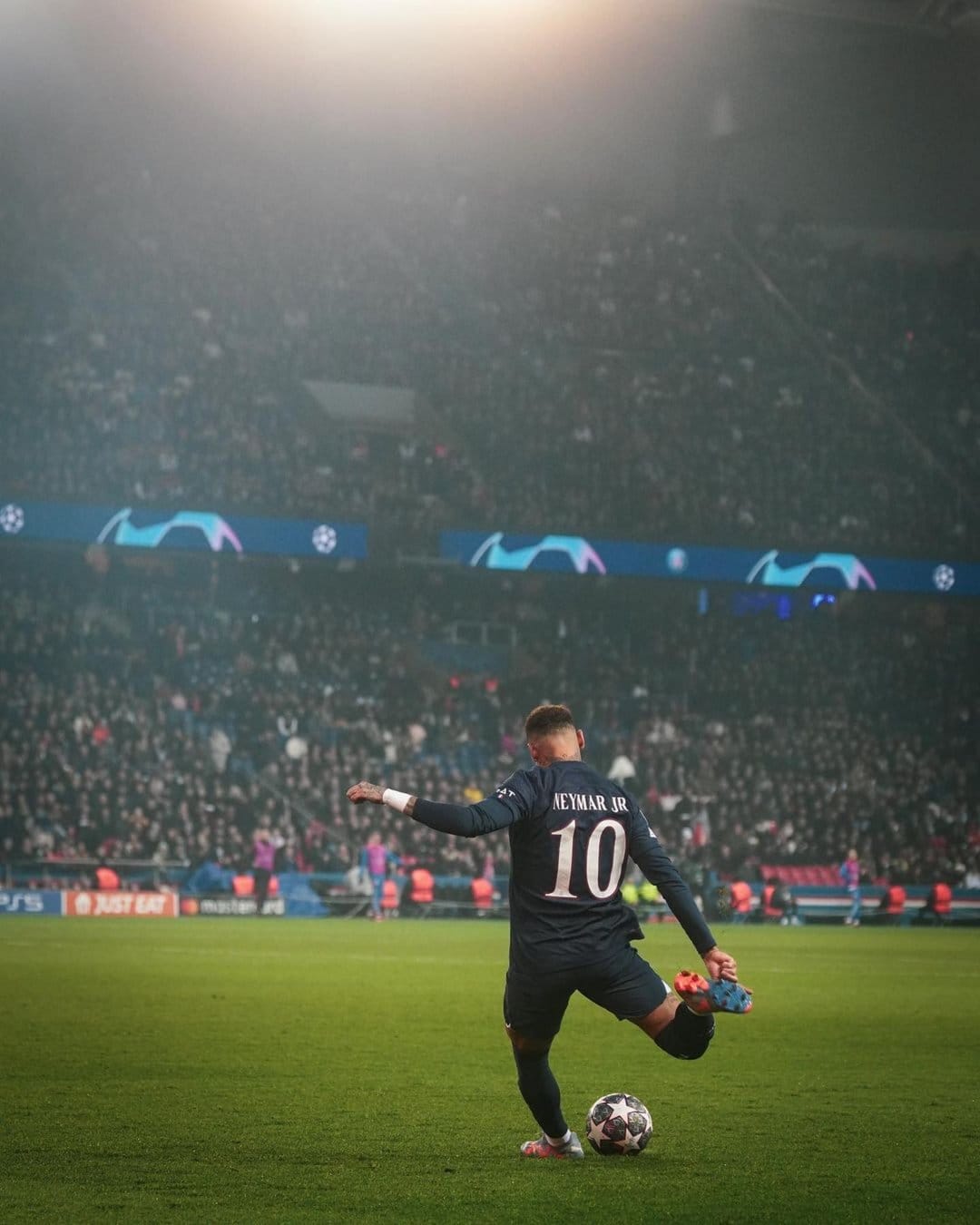 Neymar Jr wearing jersey number 10 stands on a bright green football pitch facing a crowded stadium at night.