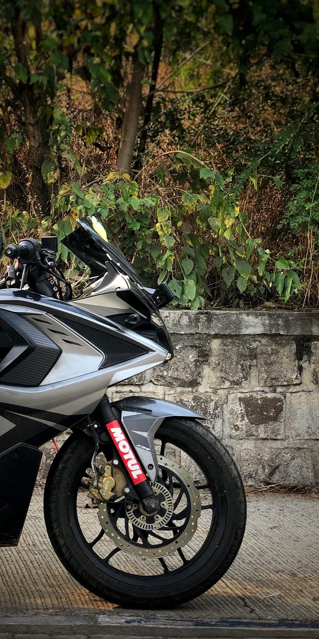 Silver sportbike front view with a tinted windshield and suspension decals parked near a stone wall.