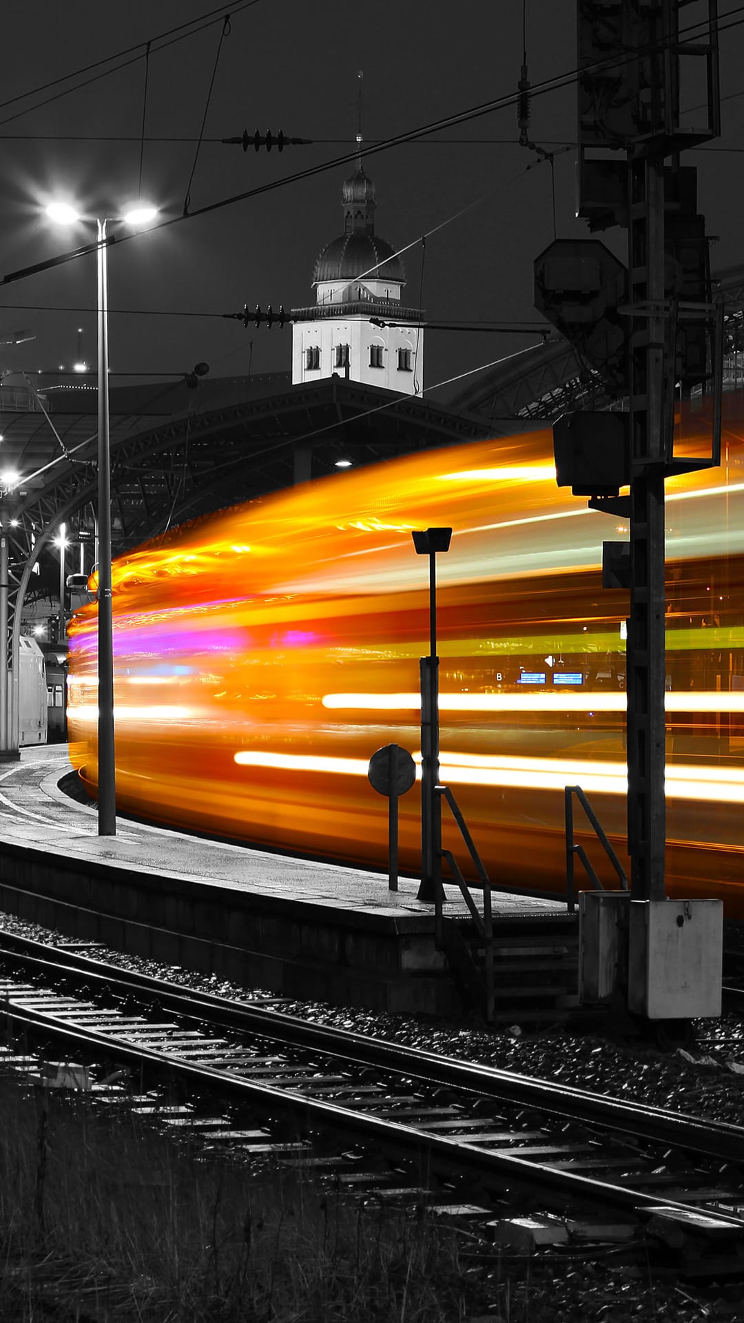 Glowing orange and purple light trails from a fast train move through a monochrome railway station platform.