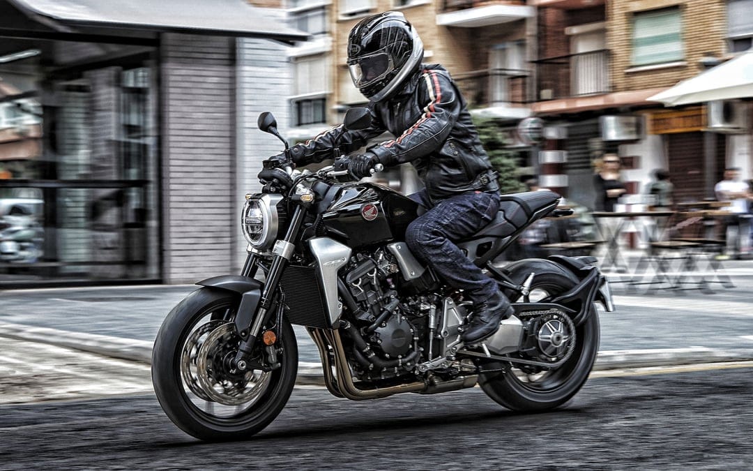 Black sport motorcycle with rider in leather gear moving through a blurred city street with outdoor cafes.