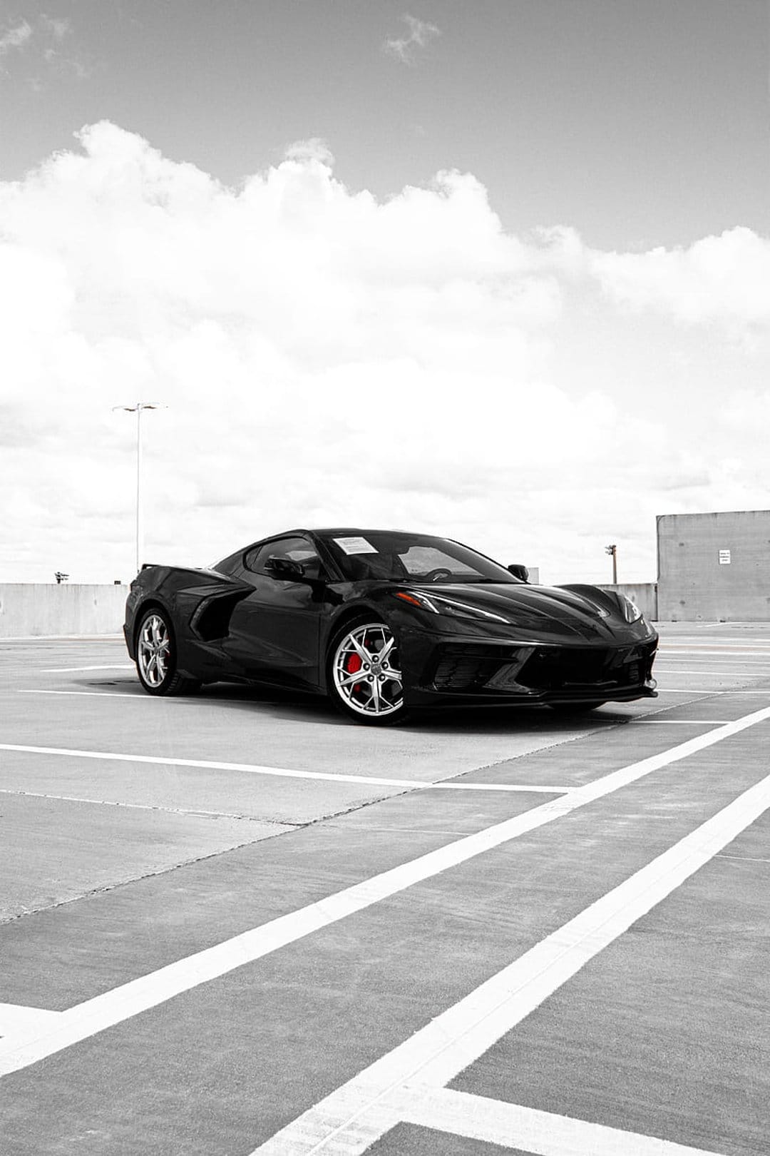 Black Chevrolet Corvette C8 parked on concrete under a bright sky with white clouds in a vertical composition.