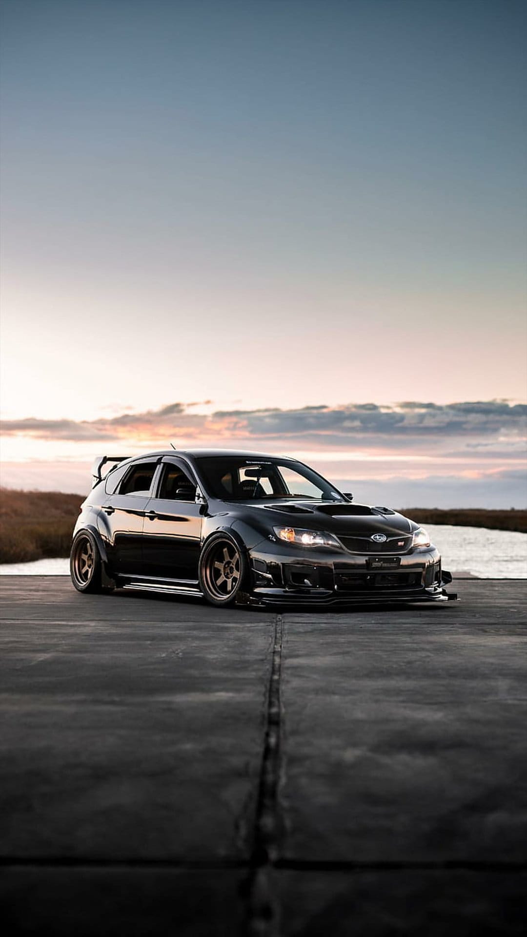 Black Subaru WRX STI hatchback with bronze wheels and a widebody kit parked on asphalt during a coastal sunset.