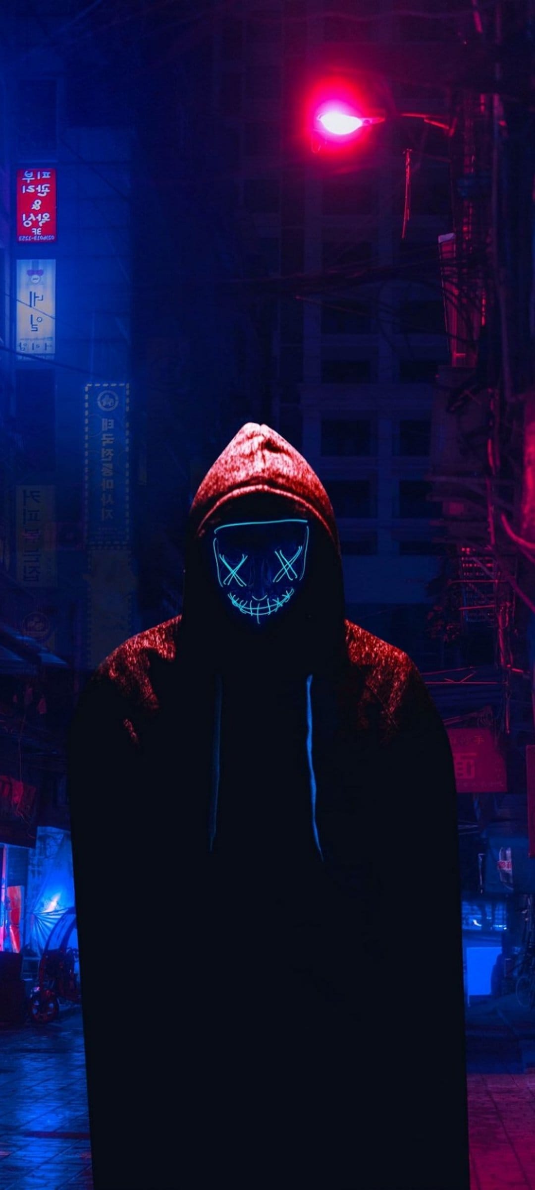 Hooded figure in a glowing neon mask stands in a dark urban alleyway illuminated by red and blue electric lights.