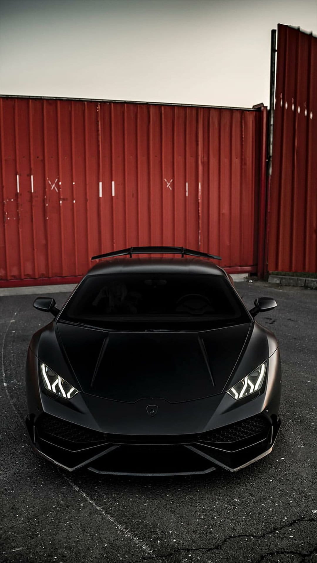 Matte black Lamborghini supercar parked in front of a red corrugated metal wall in a low-angle vertical shot.