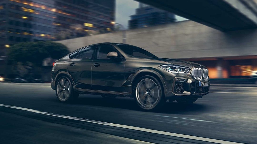 Dark metallic BMW X6 SUV driving on an urban road under a bridge with blurred city lights and glowing headlights.