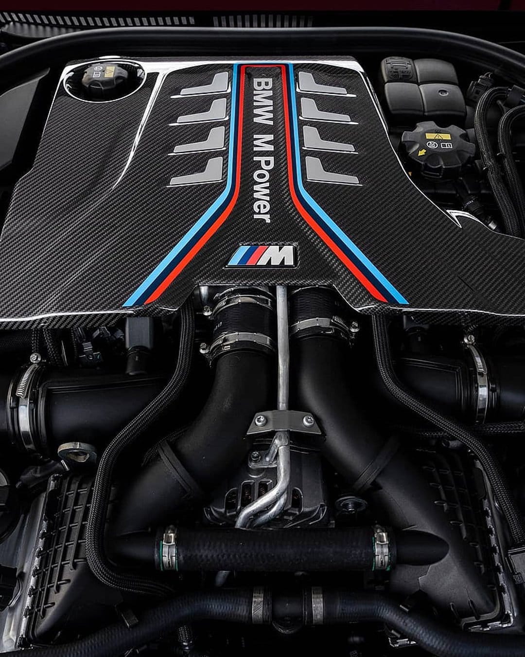 BMW M Power engine featuring a carbon fiber cover with signature colored stripes and detailed intake pipes.