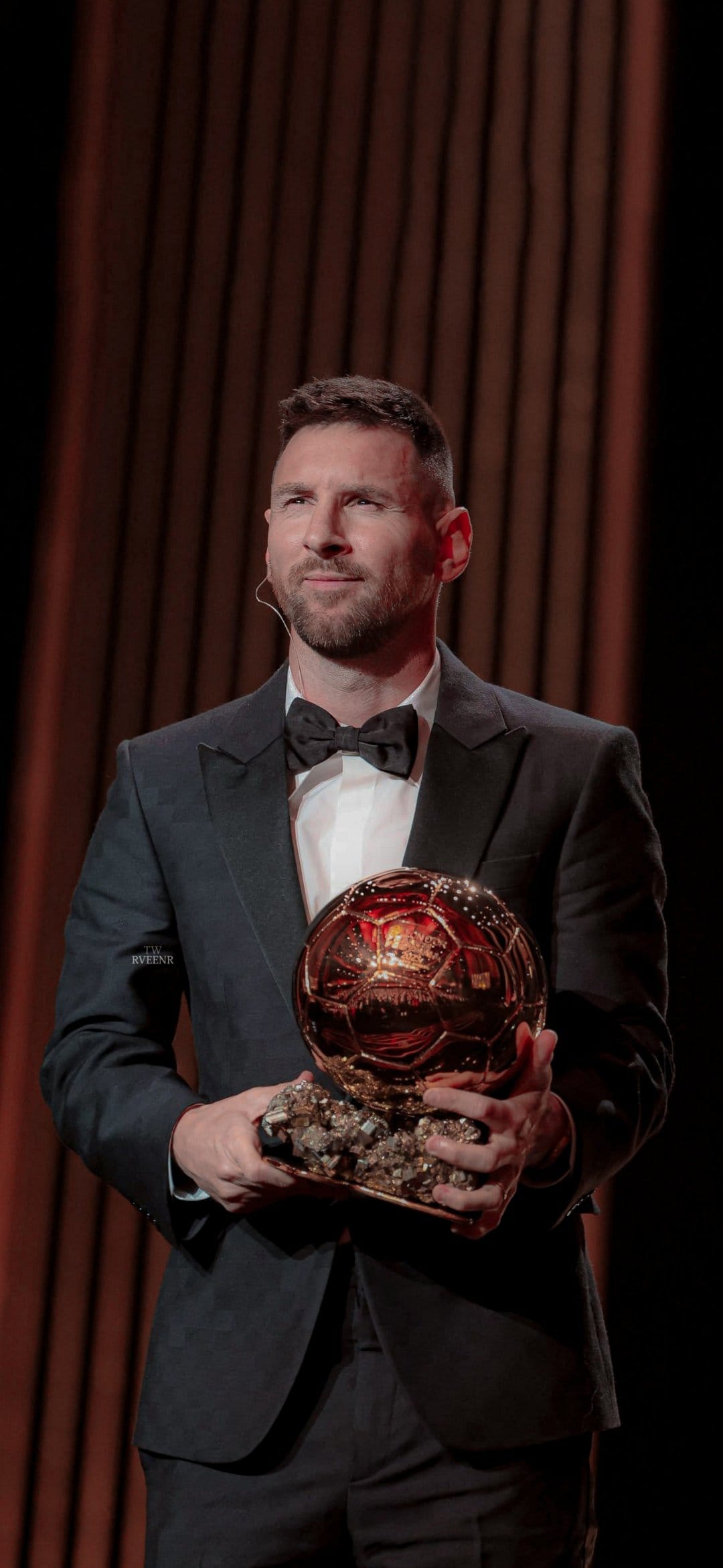 Lionel Messi in a black tuxedo holds the golden Ballon d'Or trophy while looking sideways on a dark stage.