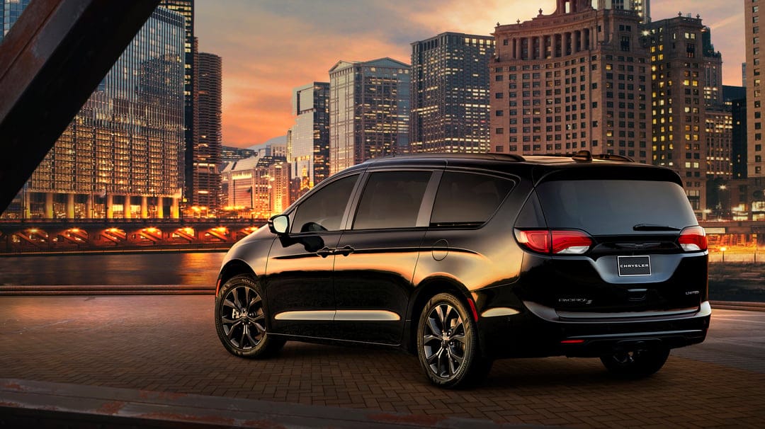 Black Chrysler Pacifica minivan parked on a brick pier with illuminated city skyscrapers during a golden hour sunset.