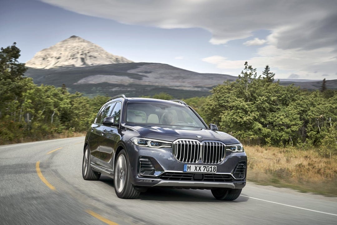 Dark grey BMW X7 SUV driving on a winding mountain road with a large pointed peak in the background.