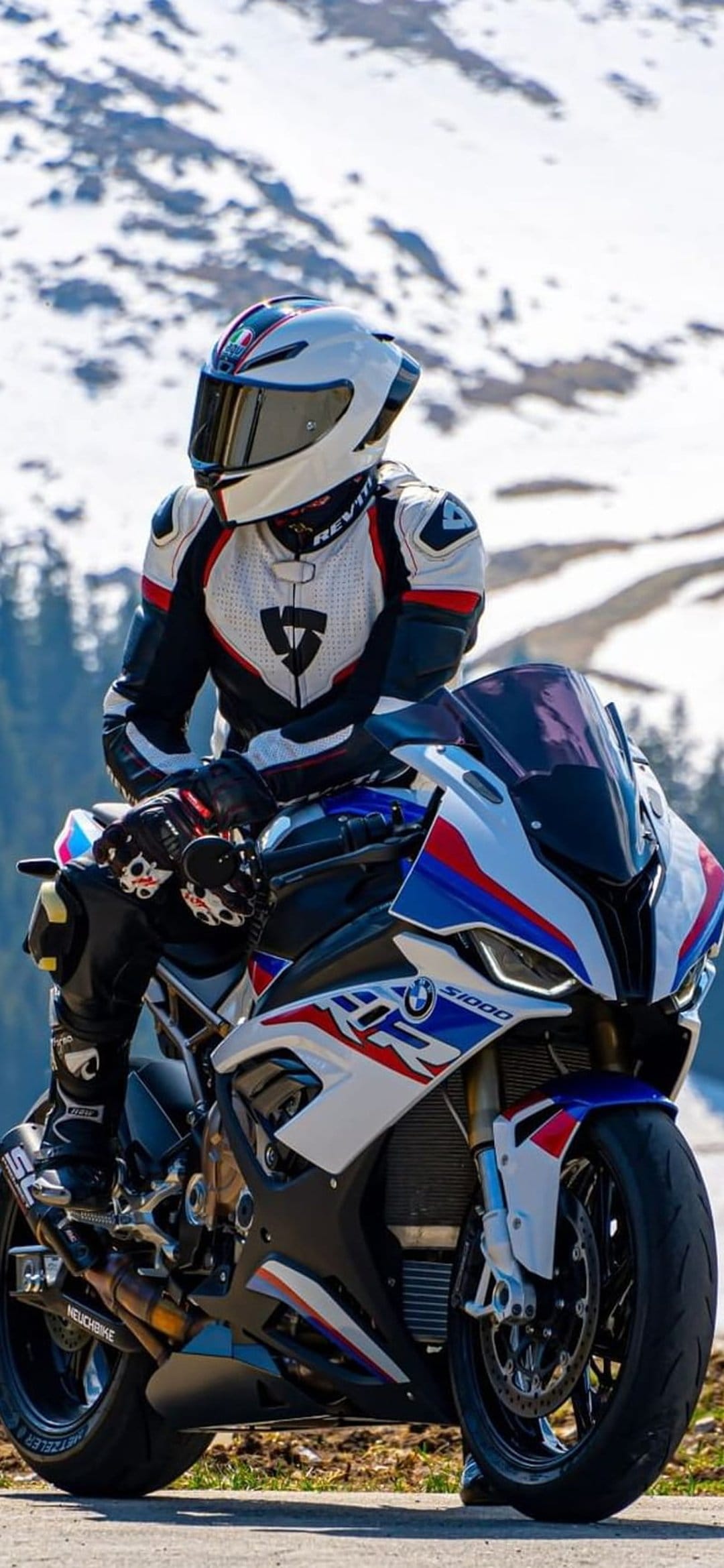 Motorcyclist in racing gear sits on a BMW S1000RR amidst a snow-covered mountain peak with distant evergreen trees.
