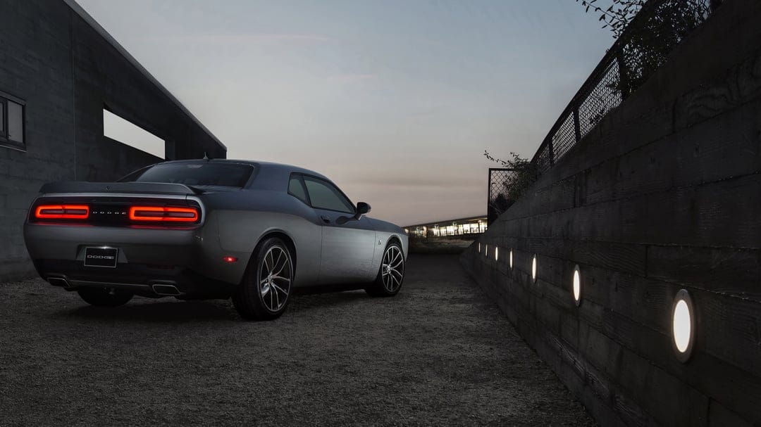 Silver Dodge Challenger with glowing red taillights parked beside a lit wooden wall at dusk on a gravel path.