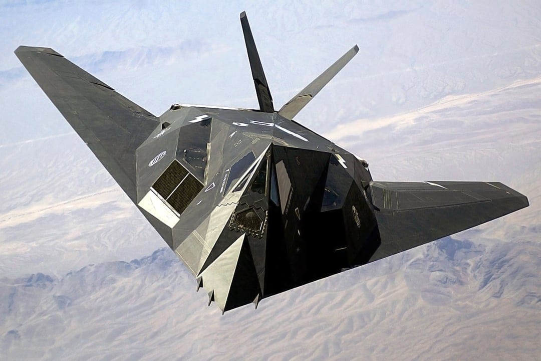 F-117 Nighthawk stealth fighter jet flying above brown desert mountains under a clear blue sky during the day.