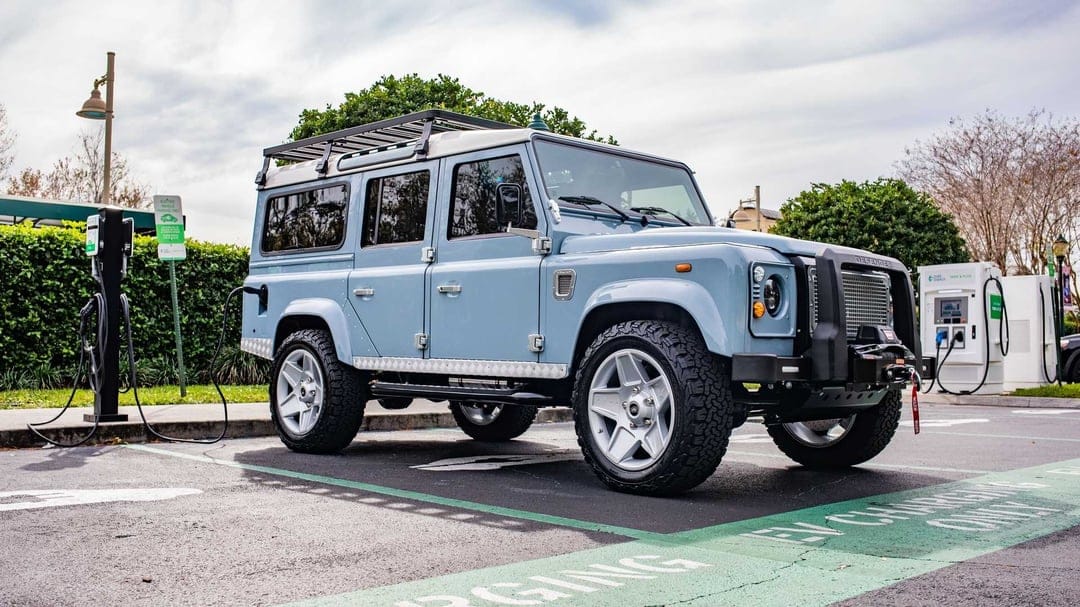 Light blue classic Land Rover Defender plugged into an EV charging station in a parking lot under soft daylight.
