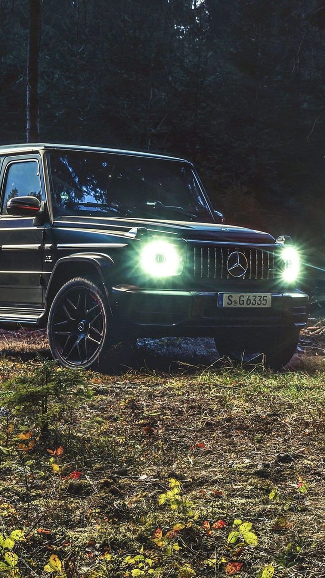Mercedes-AMG G 63 SUV with glowing headlights parked on a leafy forest floor in dim nocturnal light.