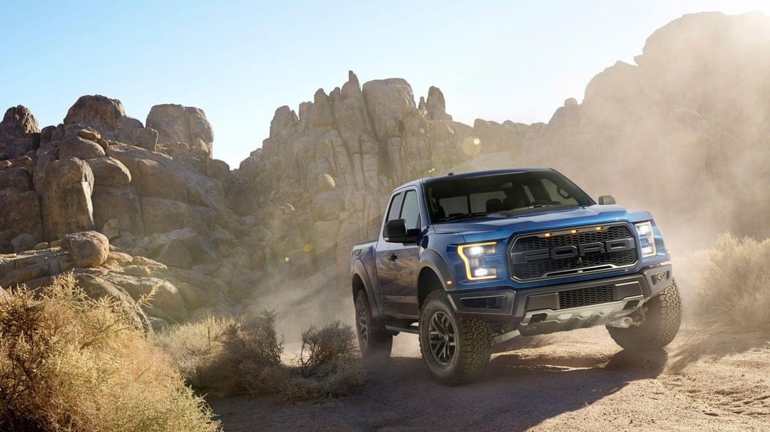 Blue Ford Raptor truck driving through a dusty desert with massive rock formations and bright golden sunlight.