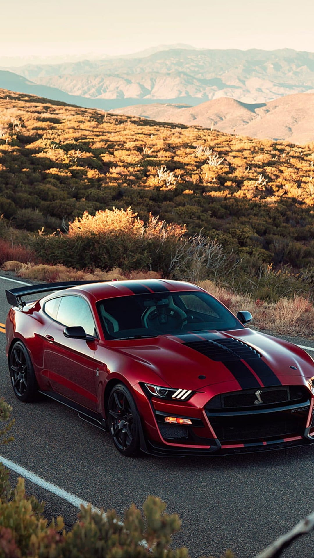 Red Ford Mustang Shelby GT500 with black stripes driving on a paved road through arid desert mountain peaks.