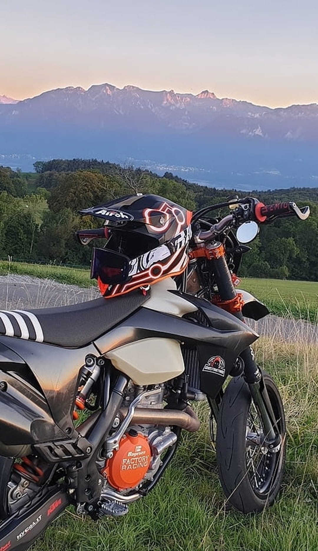 Orange dirt bike with a helmet on the seat overlooks jagged mountain peaks under a glowing orange and purple sunset.