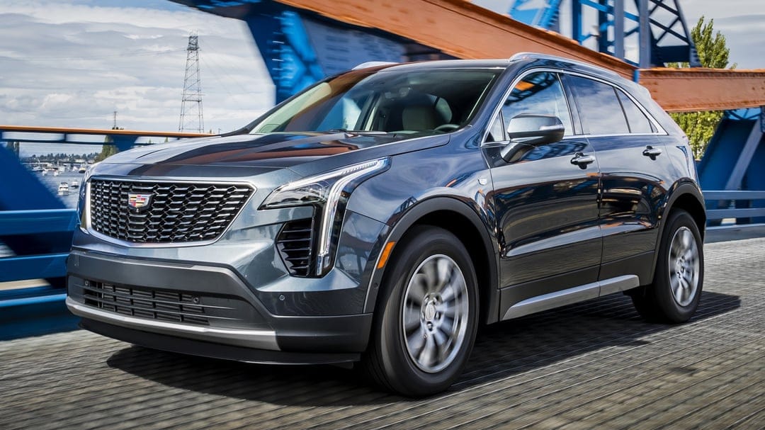 Dark grey luxury SUV driving on a blue and orange bridge with motion-blurred wheels and scenic city backdrop.