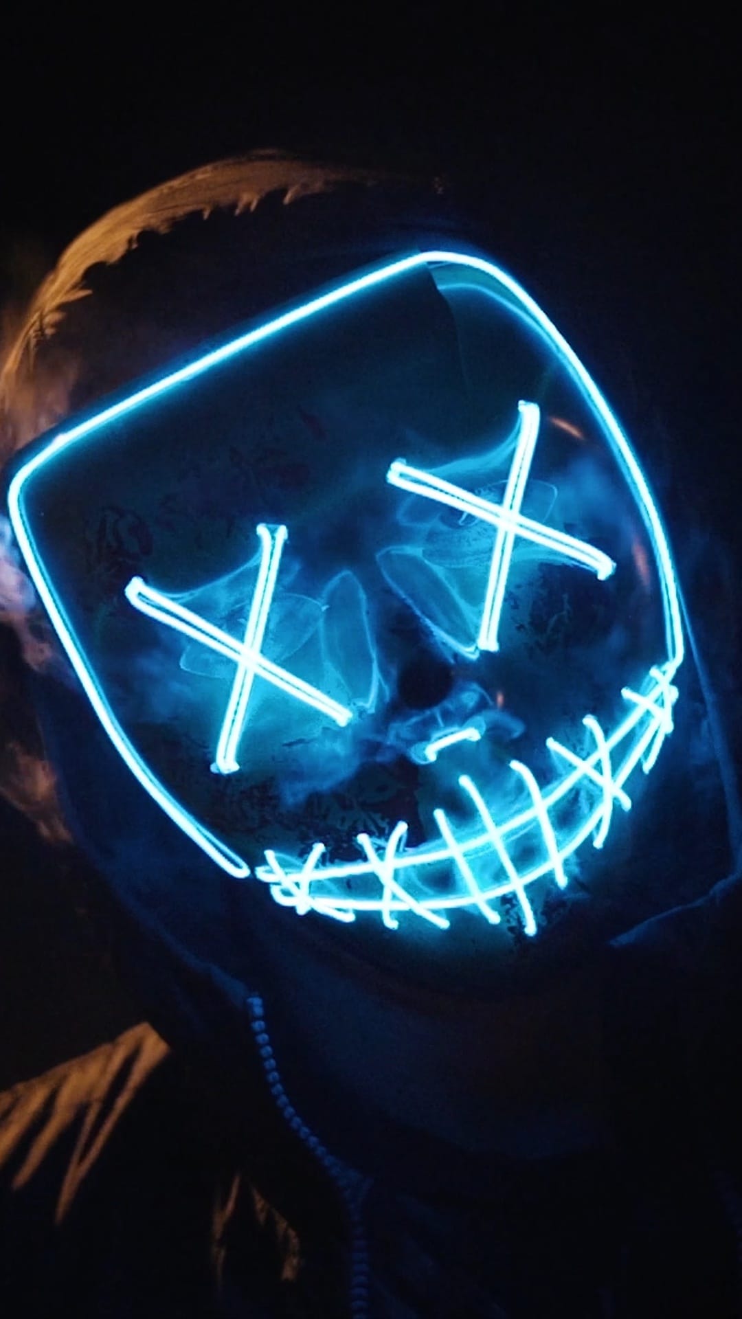 Person wearing a black mask with blue neon X-shaped eyes and a glowing smile surrounded by thin white vapor.