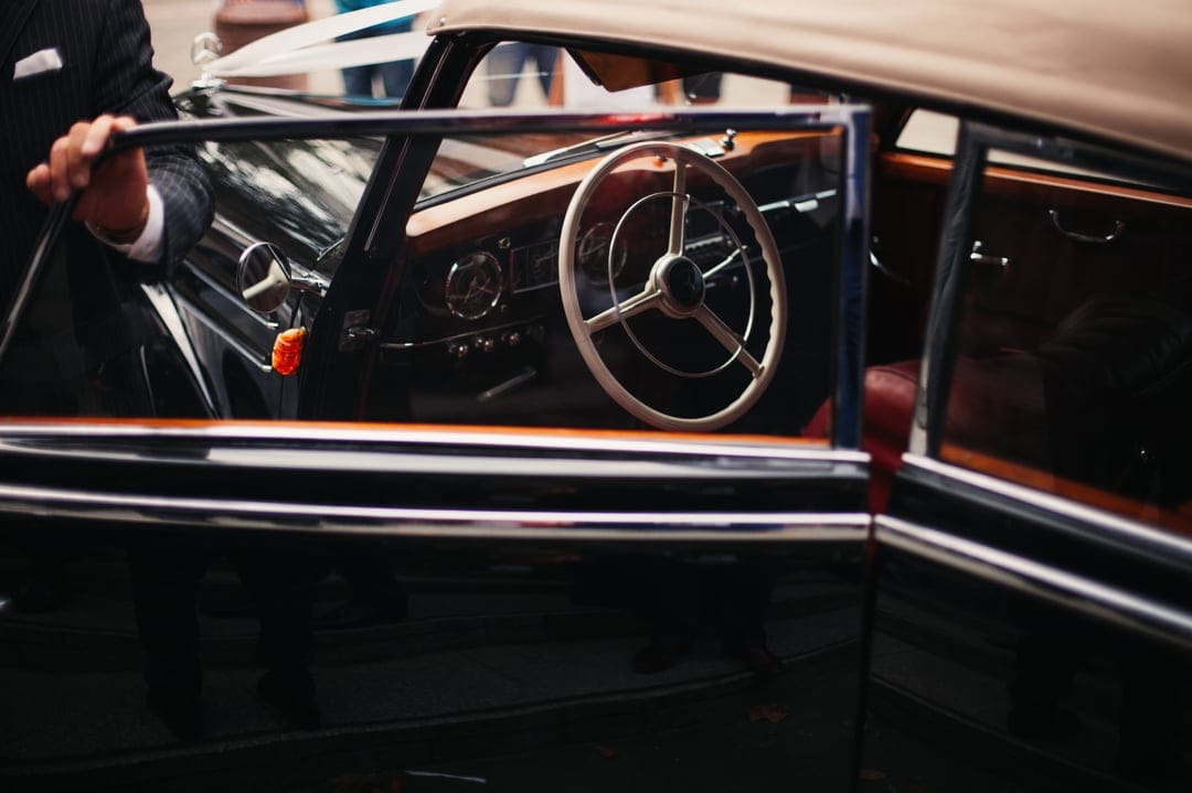 Man in a dark suit holding the door of a vintage car with a wooden dashboard and red leather upholstery.