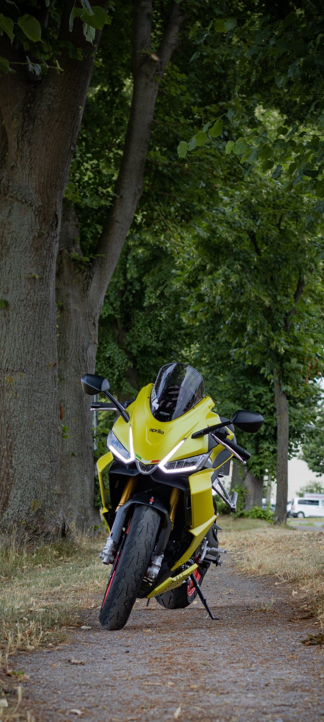 Yellow Aprilia motorcycle with glowing LED lights parked on a woodland dirt path surrounded by tall green trees.