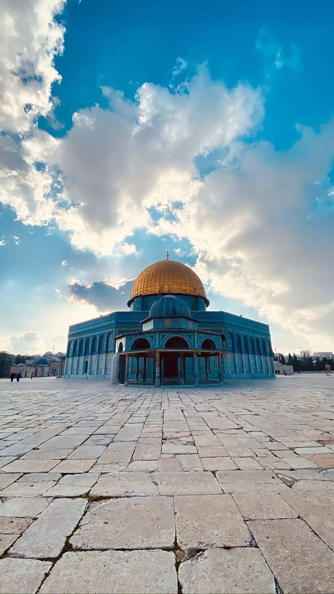 Golden Dome of the Rock centered on a stone courtyard under a dramatic sky with sunbeams piercing through clouds.