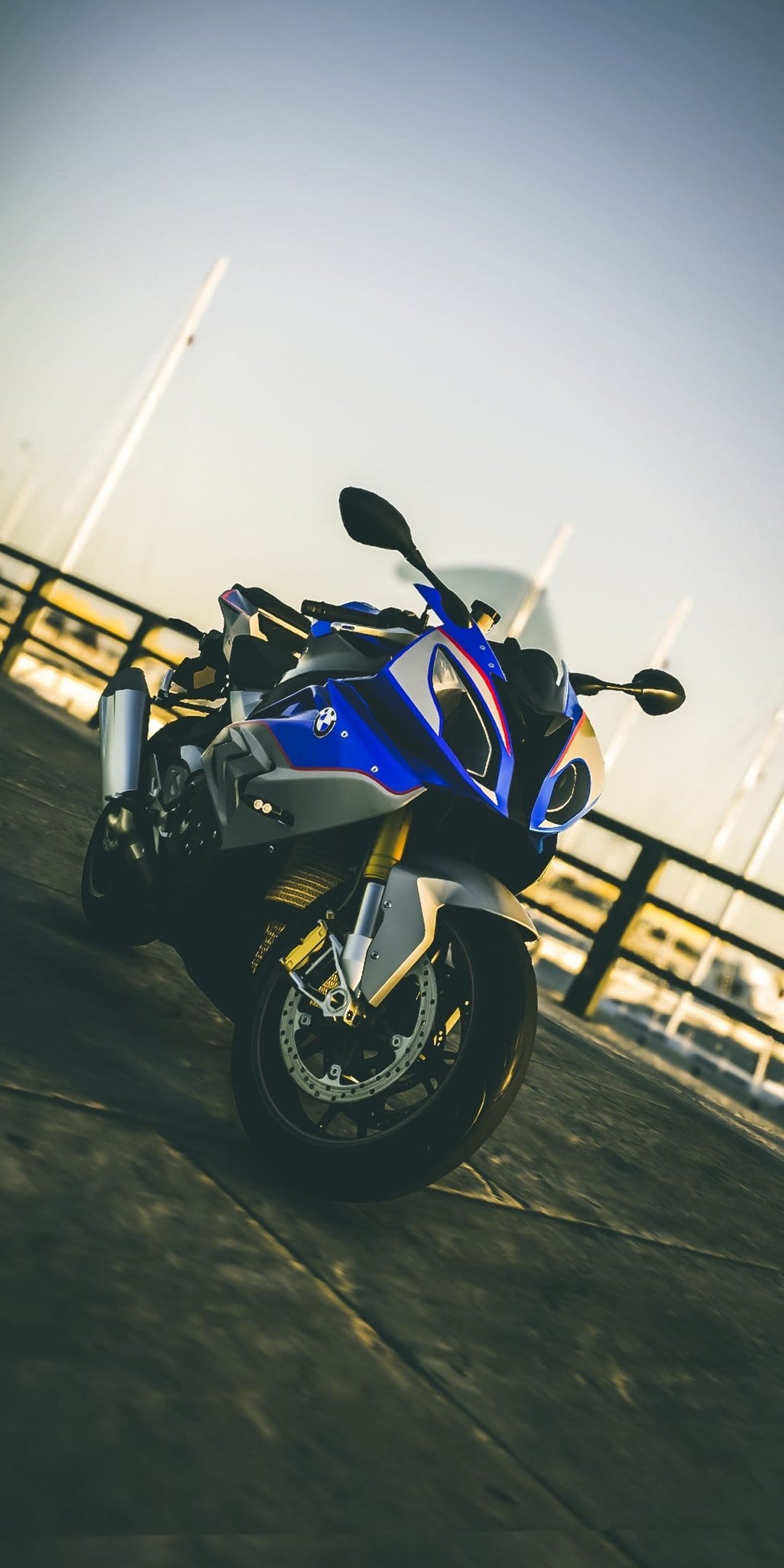 BMW S1000RR motorcycle in blue and white parked on a wooden dock with sunset lighting and boat masts behind it.