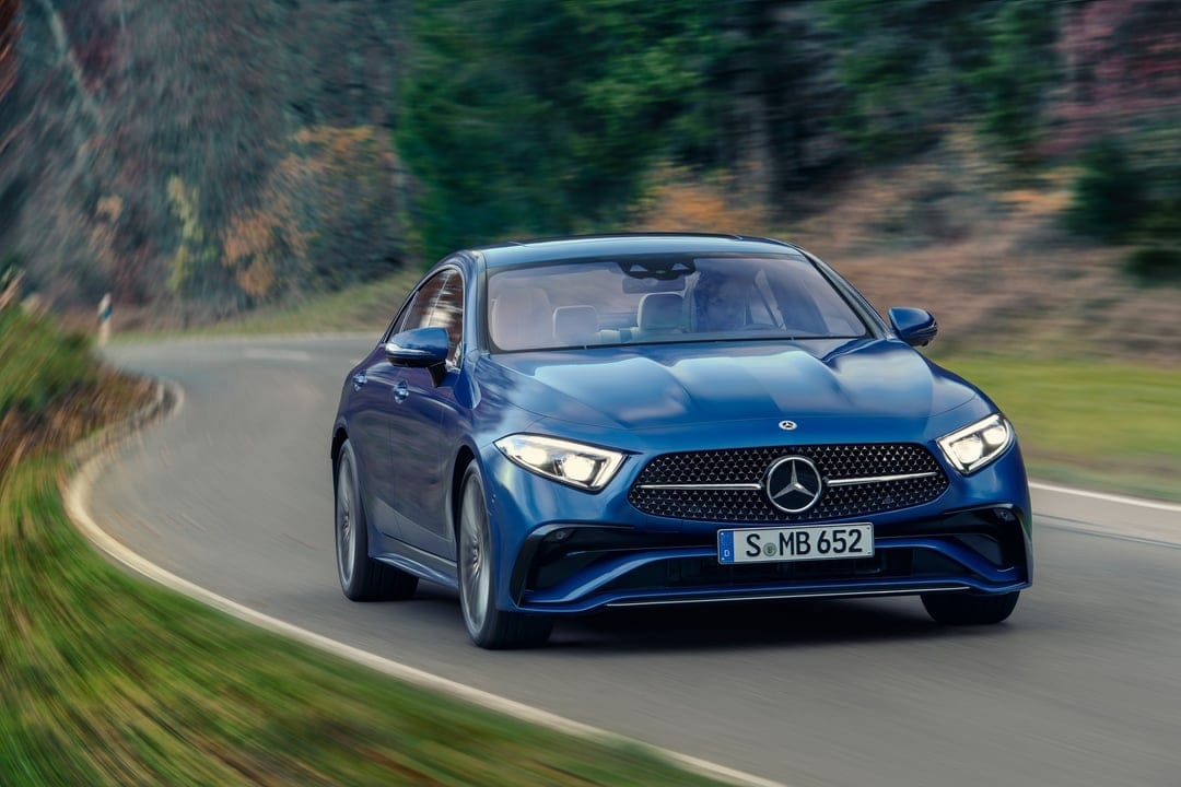 Blue Mercedes-Benz luxury car driving on a curved road through a blurred forest with headlights on.