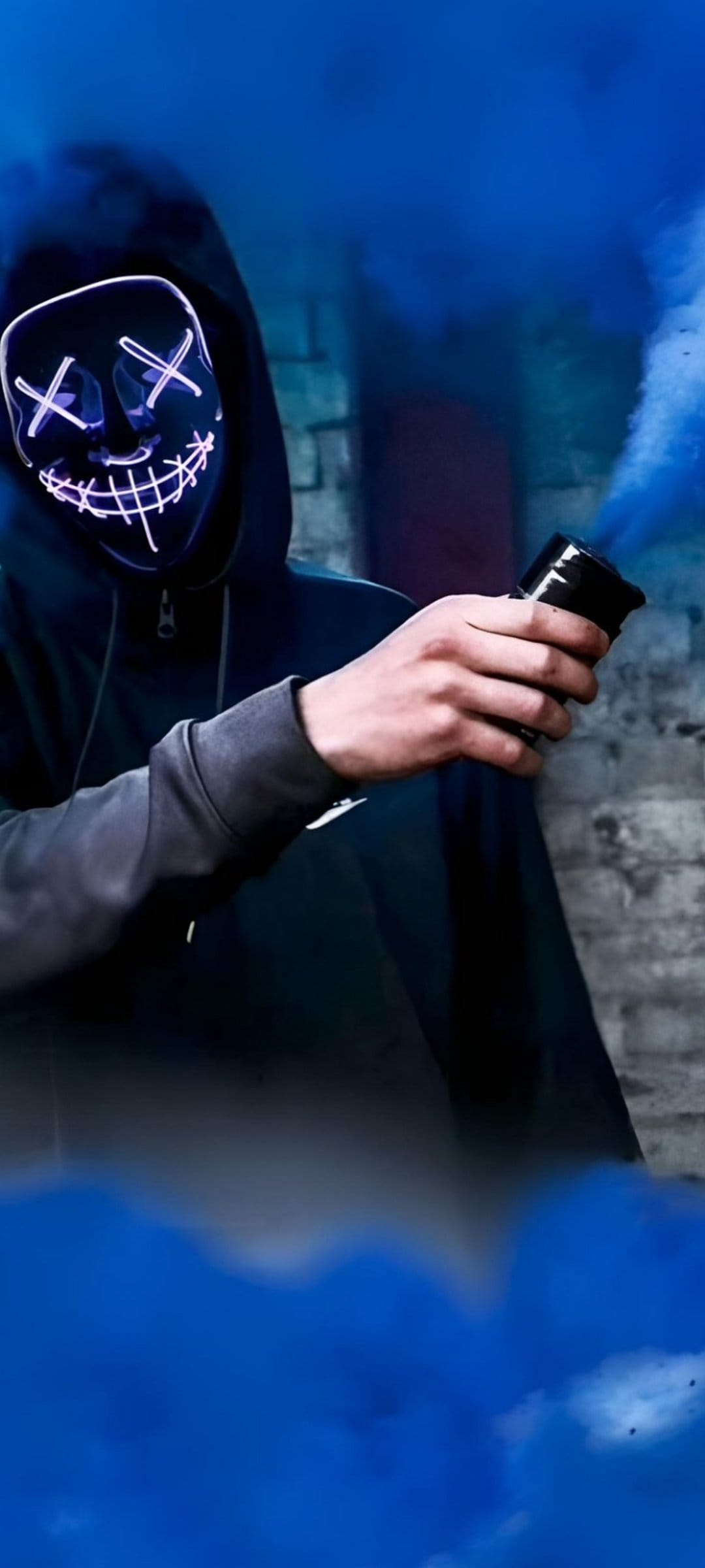 Person wearing a glowing blue LED mask holds a smoke bomb with thick azure clouds against a dark brick wall.