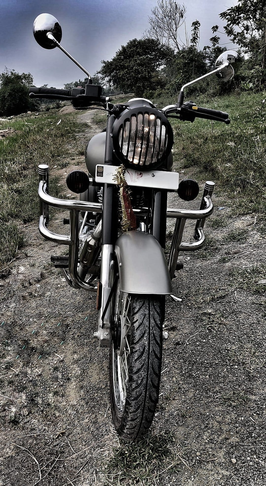 Classic motorcycle front view with a grilled headlight and chrome bars parked on a grassy field under grey clouds.