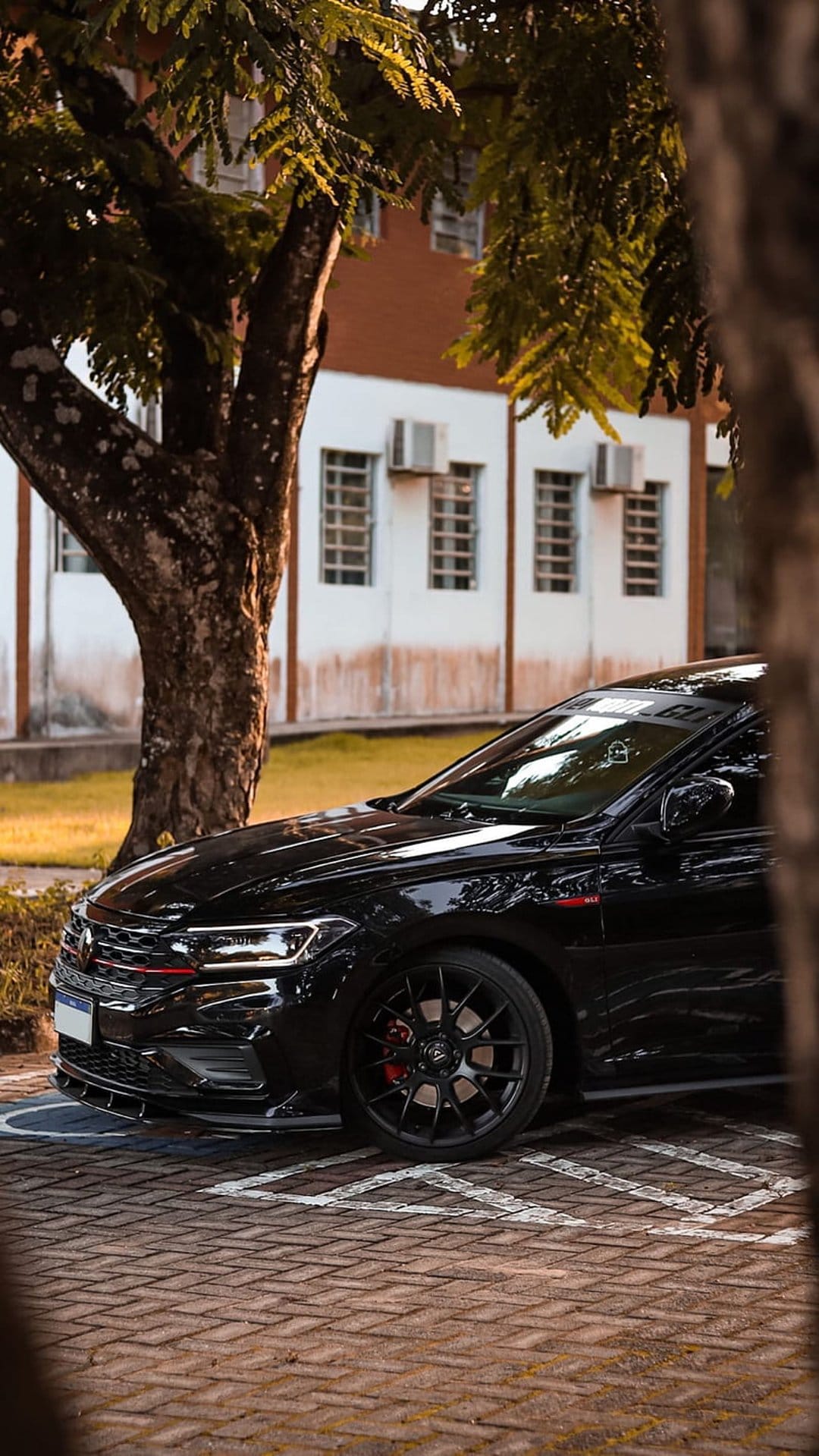 Black sedan with red brake calipers parked on a brick lot under a leafy tree near a modern white building.