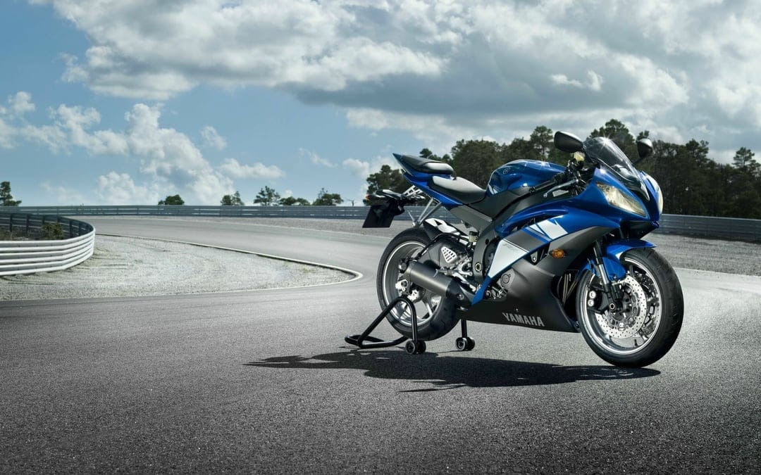 Blue Yamaha YZF-R6 motorcycle on a rear stand on a curved asphalt race track under a bright cloudy sky.
