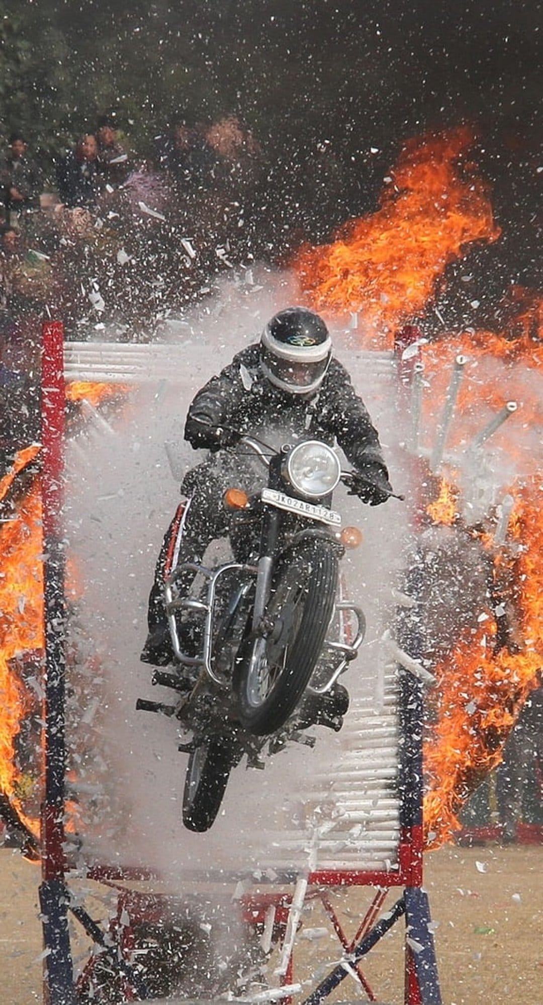 Motorcyclist in protective gear leaps through a wall of burning wood splinters and bright orange fire in mid-air.