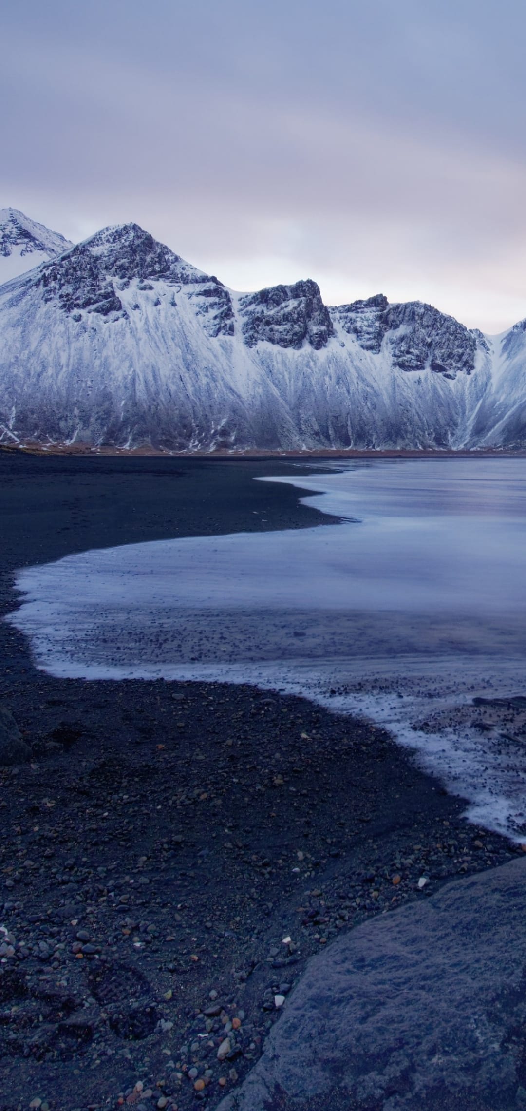 Snow-covered mountains rise above a dark volcanic beach and partially frozen glacial water under a grey sky.