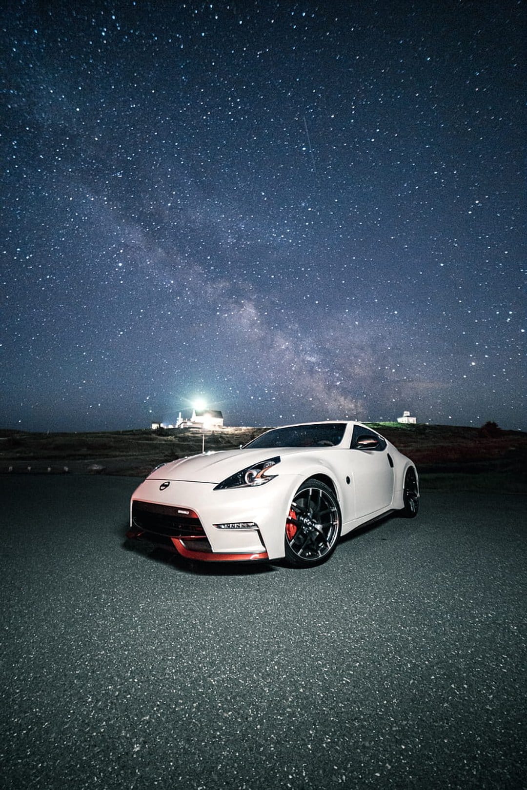 White Nissan 370Z sports car parked on an asphalt road at night under a star-filled Milky Way galaxy sky.