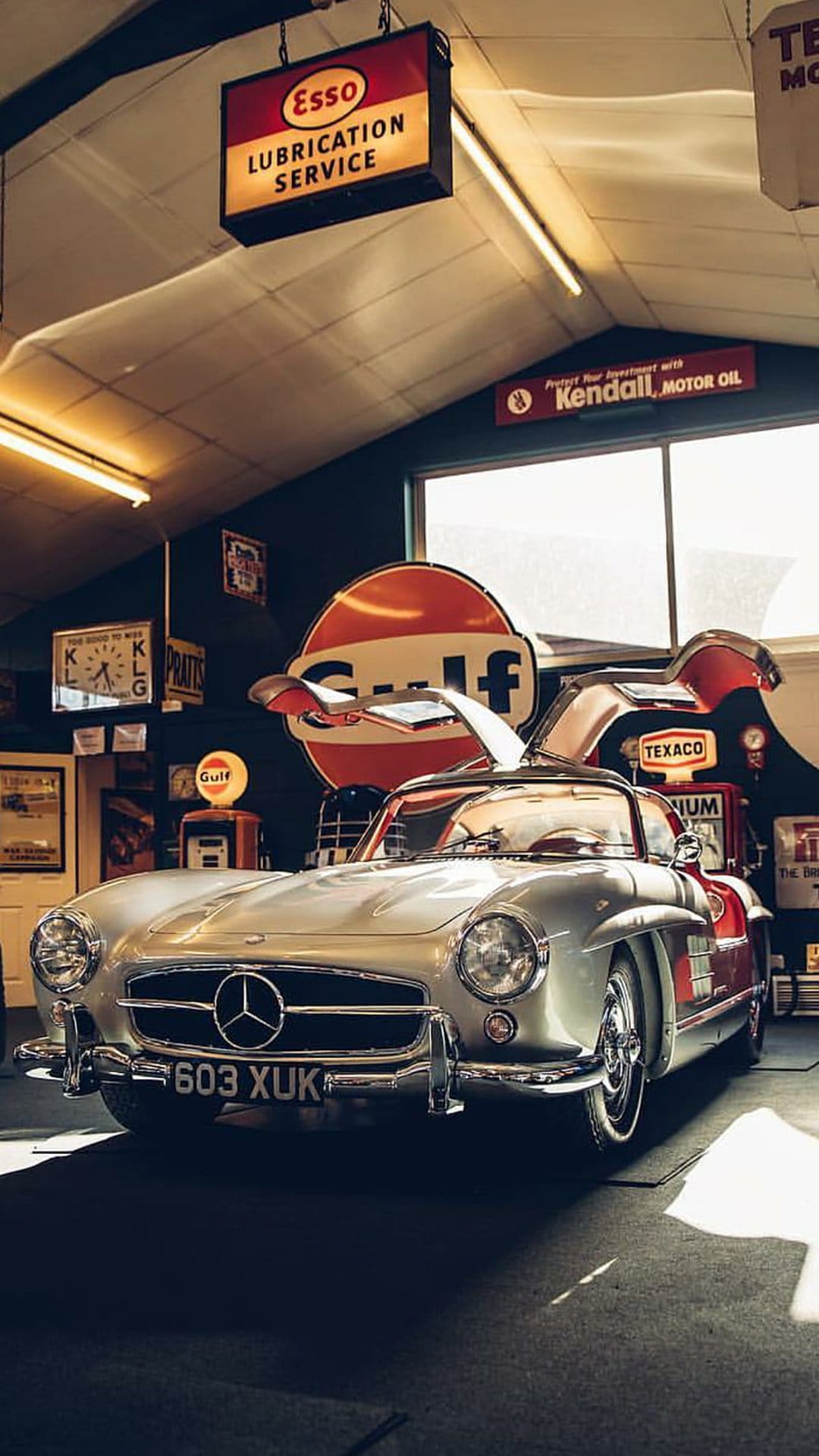 Mercedes-Benz 300 SL Gullwing with open doors in a garage filled with vintage Esso and Gulf gasoline signs.