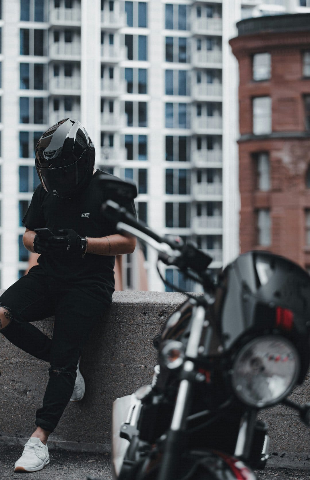Motorcyclist in a black helmet sits on a stone ledge checking a phone with a blurred bike in the foreground.