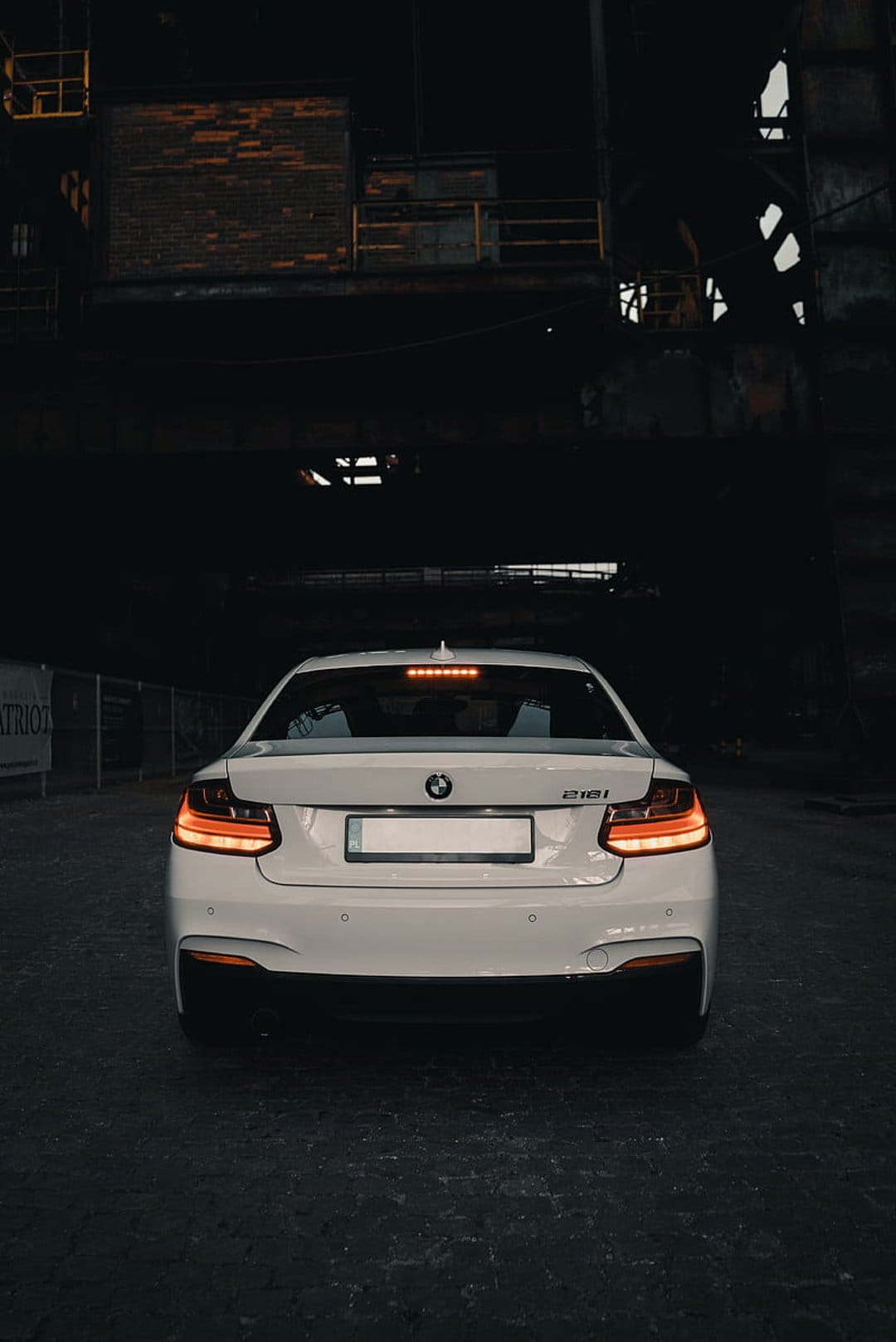 White BMW 218i coupe rear view with red taillights in a dark warehouse with brick walls and metal beams.