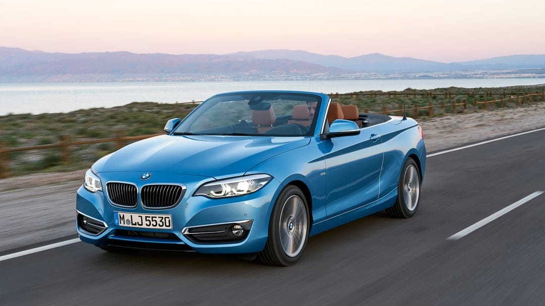 Blue BMW 2 Series Convertible driving on a coastal road with mountains and water under a soft sunset sky.