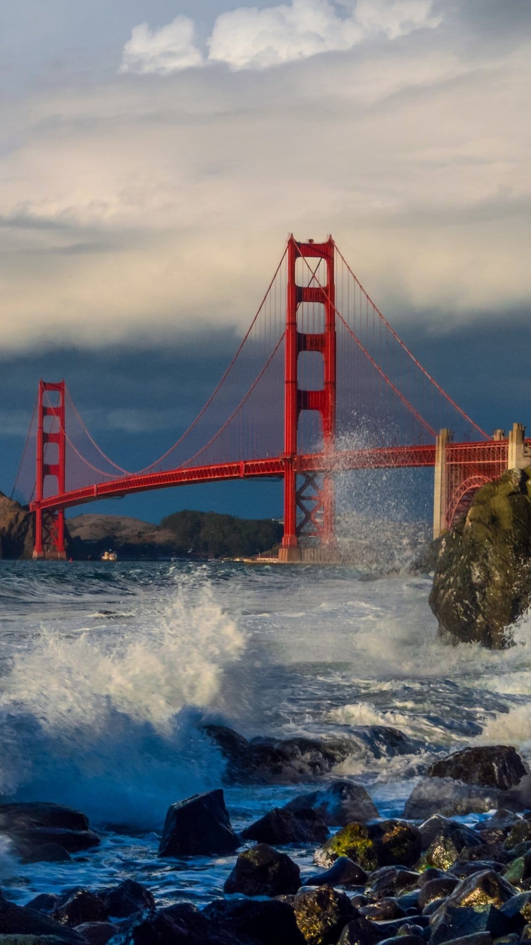 Golden Gate Bridge stands above crashing white ocean spray and dark rocks under a heavy grey overcast sky.