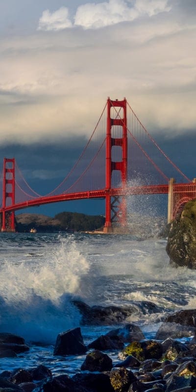 This dynamic image captures the iconic Golden Gate Bridge, its vibrant red structure standing boldly against a dramatic, cloudy sky. Powerful ocean waves crash against rugged rocks in the foreground, creating spectacular white spray that emphasizes the raw energy of the sea. The scene offers a breathtaking view of this architectural marvel amidst the powerful forces of nature.