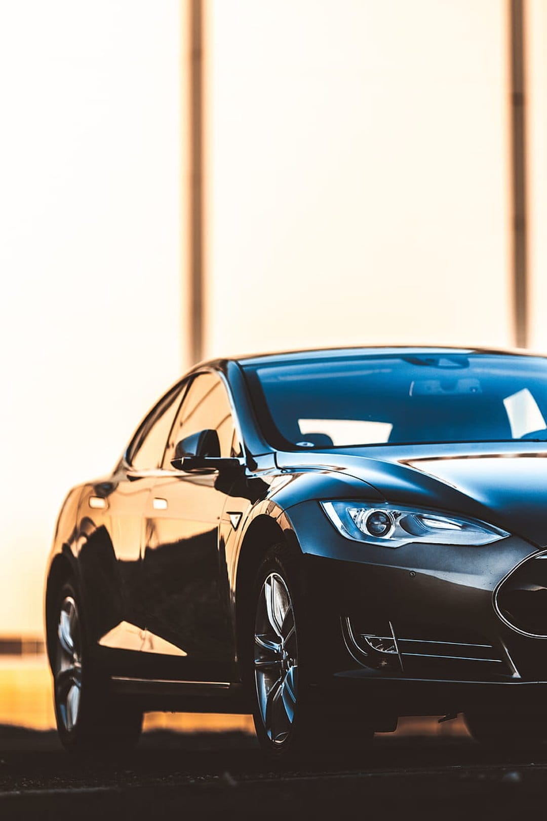Black Tesla Model S headlight and front fender reflecting warm golden hour light against a blurred background.