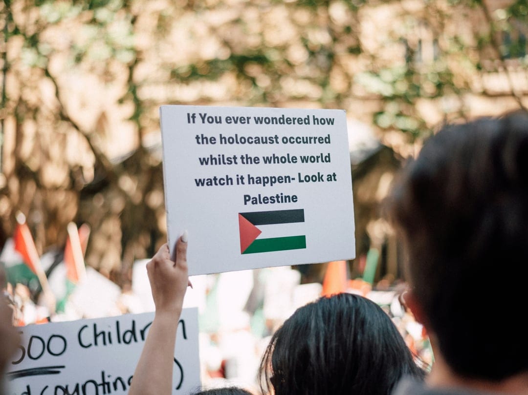 Protest sign held by hands features text about Palestine and the Palestinian flag at a public demonstration.